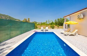 En rektangulär pool med klart, blått vatten, omgiven av en terrass med kakelplattor. Där finns två vita solstolar med randiga kuddar och ett gult parasoll. Ett grönt staket omger området, och i bakgrunden syns en byggnad med en vit fasad och ett rött tak.