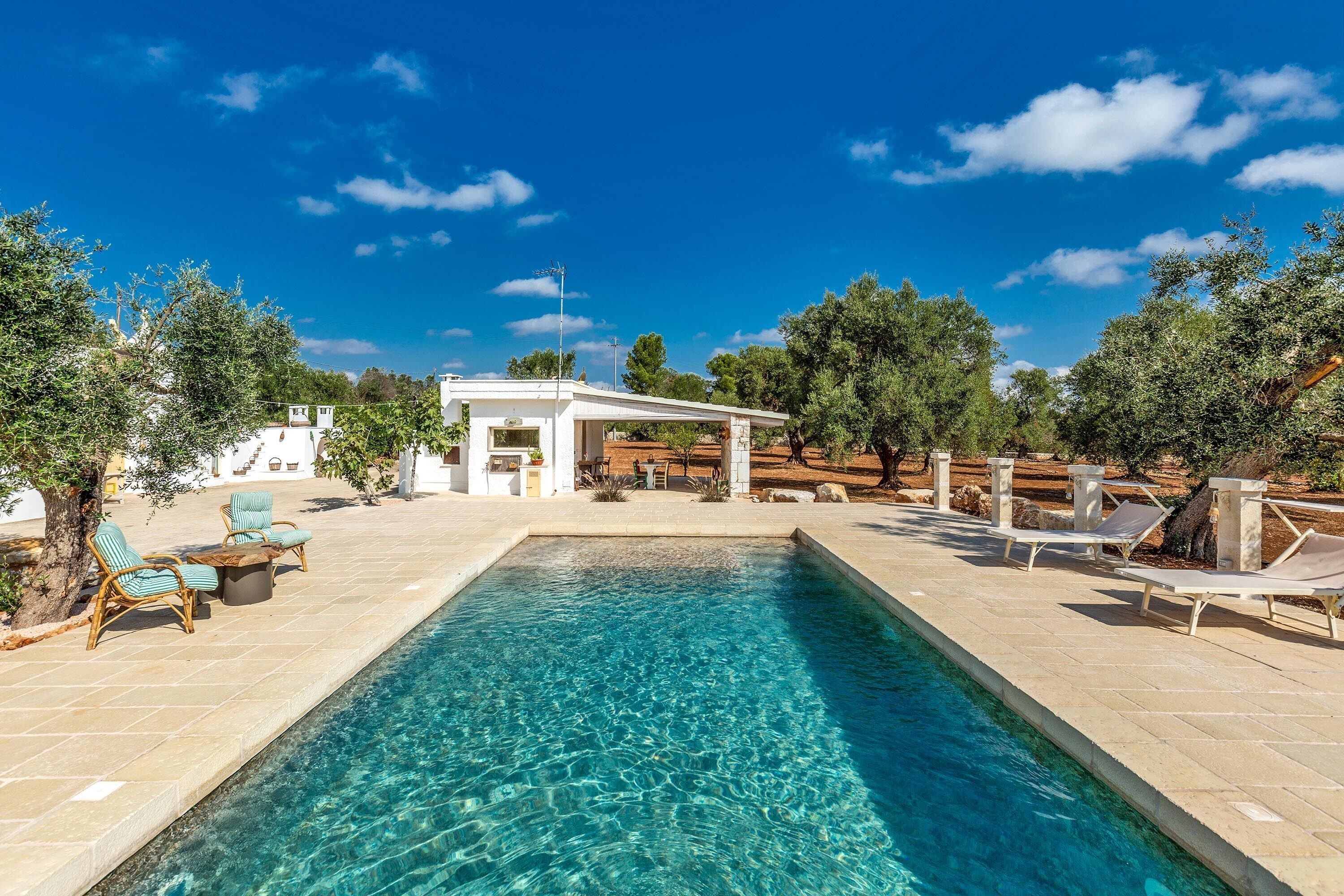 A rectangular pool with clear blue water, surrounded by lounge chairs and a covered seating area.