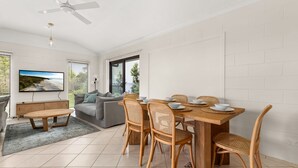 A modern living room with a sofa, a dining table with chairs, a TV mounted on the wall, and a ceiling fan.
