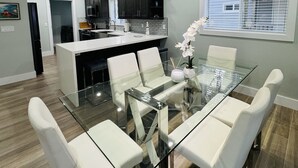 Een moderne keuken met donkere kastjes, een centraal eiland en een eethoek met een glazen tafel en witte stoelen.