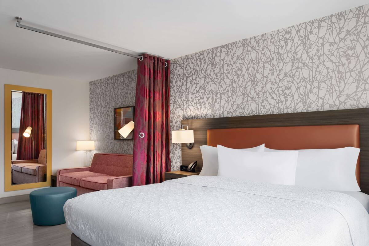 Ein Hotelzimmer mit einem großen Bett, einem rot gepolsterten Sessel, einem blauen Hocker, einem Spiegel und einer Wand mit floralem Muster.