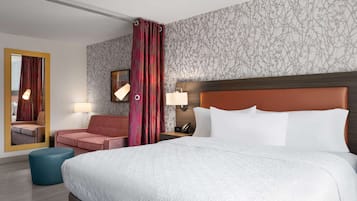 A hotel room with a large bed, a red upholstered chair, a blue ottoman, a mirror, and a wall with a floral pattern.