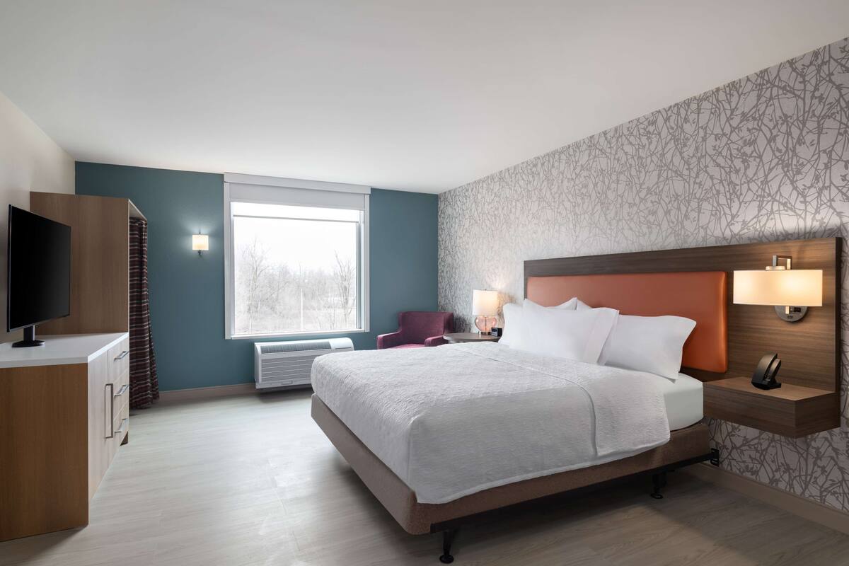 Ein modernes Hotelzimmer mit einem großen Bett, einem Flachbildfernseher, einem Schreibtisch und einem Fenster mit Blick auf Bäume.