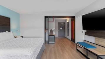 A modern hotel room with a large bed, a desk, a bench, and a flat-screen TV.