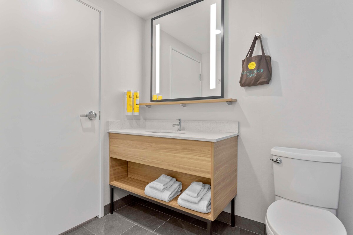 Een moderne badkamer met een witte wasbak, houten opbergmeubel en een toilet. Een spiegel met verlichting hangt boven de wasbak, en er ligt een handdoek op het opbergmeubel.