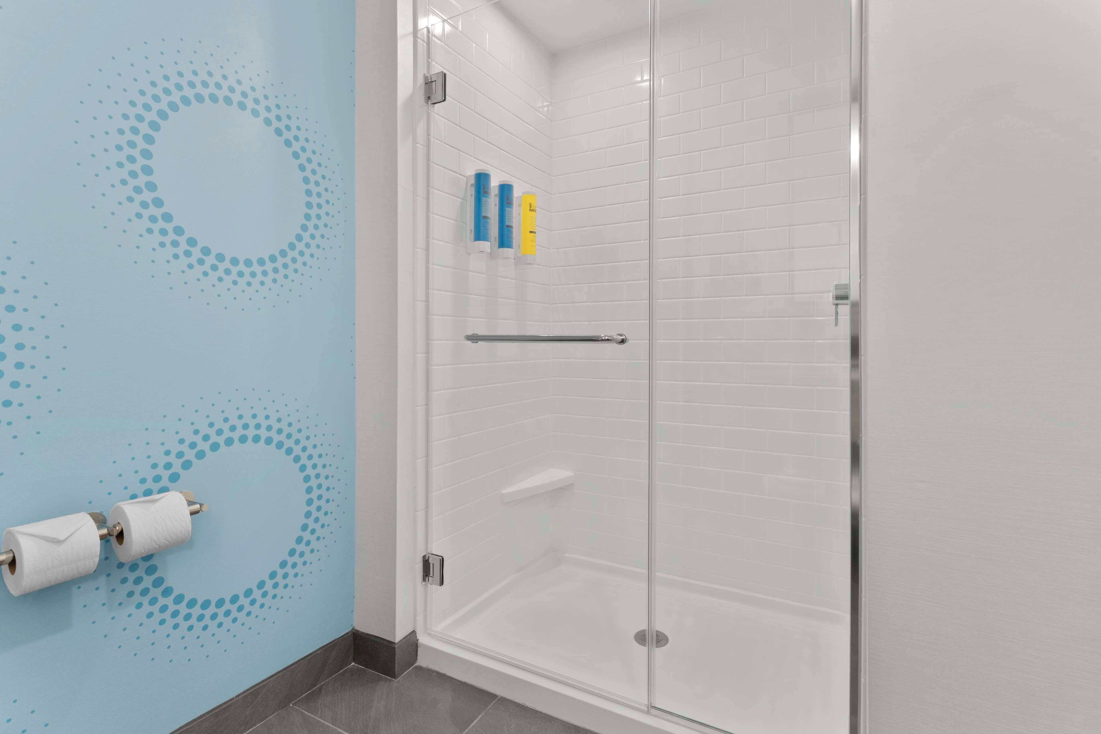 Une salle de bain moderne avec une douche à l’italienne en verre, des murs recouverts de carreaux blancs et un mur bleu orné d’un motif circulaire.