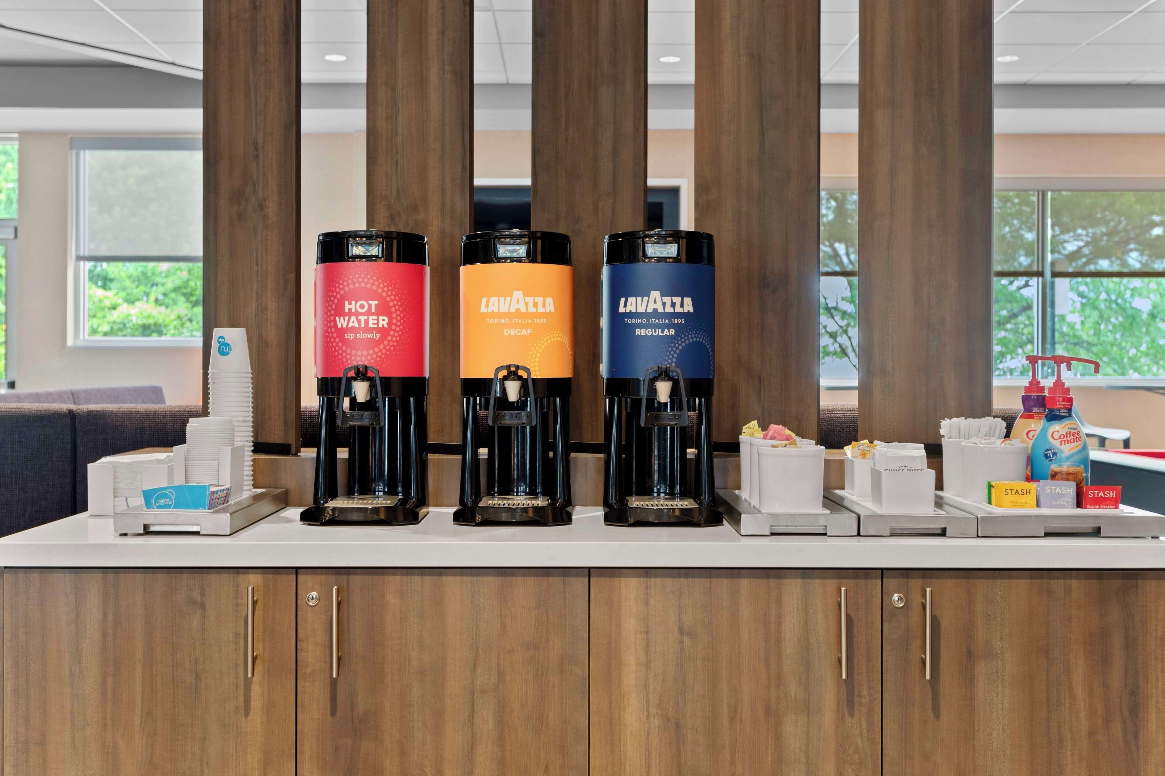 Een koffiehoek met drie koffiezetapparaten, voorzien van de aanduidingen 'LAVATTA' en 'HEET WATER', in een moderne omgeving met houten lambrisering en grote ramen.