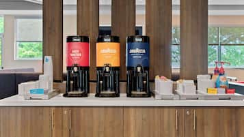 Een koffiehoek met drie koffiezetapparaten, voorzien van de aanduidingen 'LAVATTA' en 'HEET WATER', in een moderne omgeving met houten lambrisering en grote ramen.