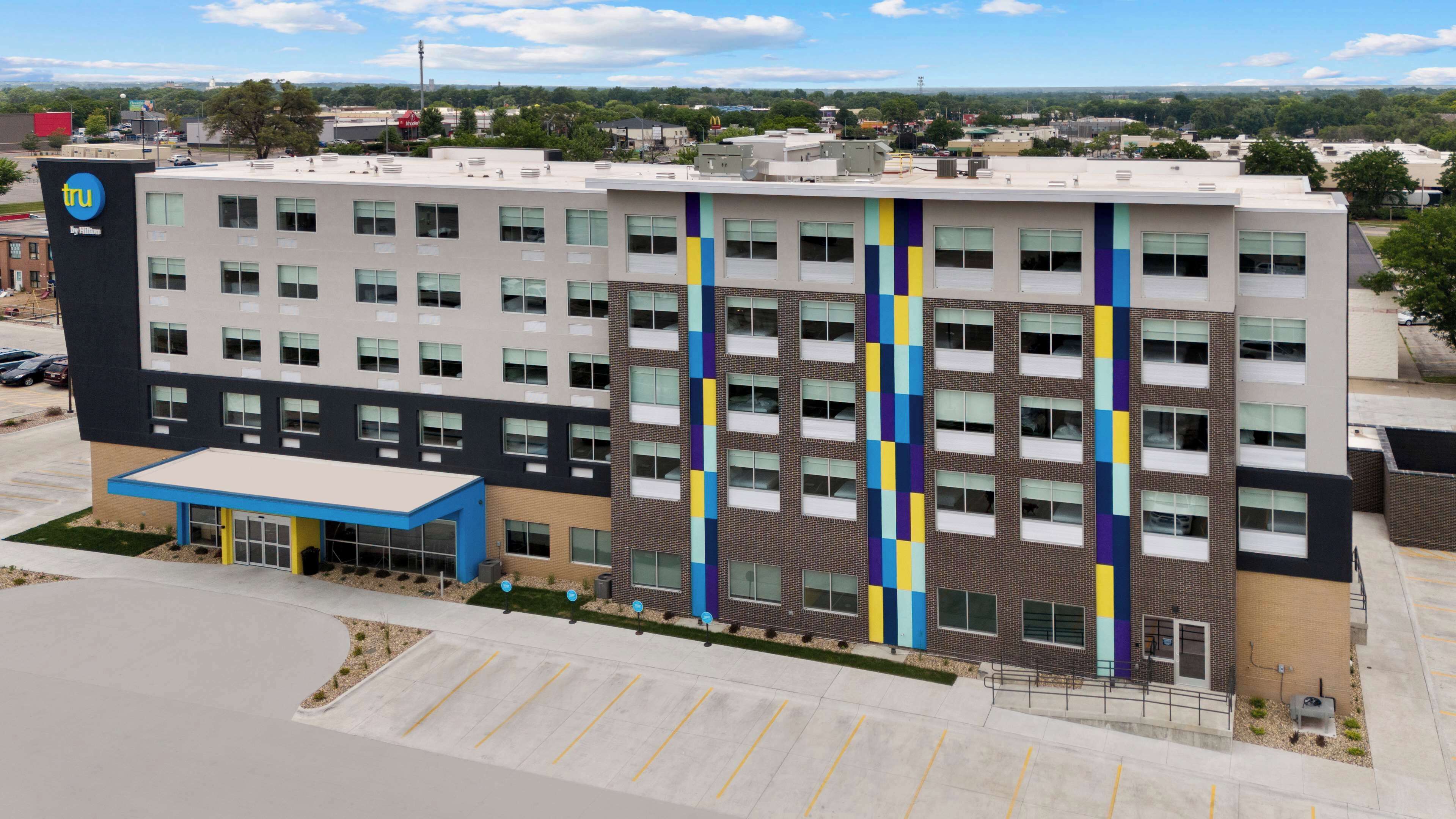 A modern hotel building with a flat roof, multiple floors, and a parking lot in front.