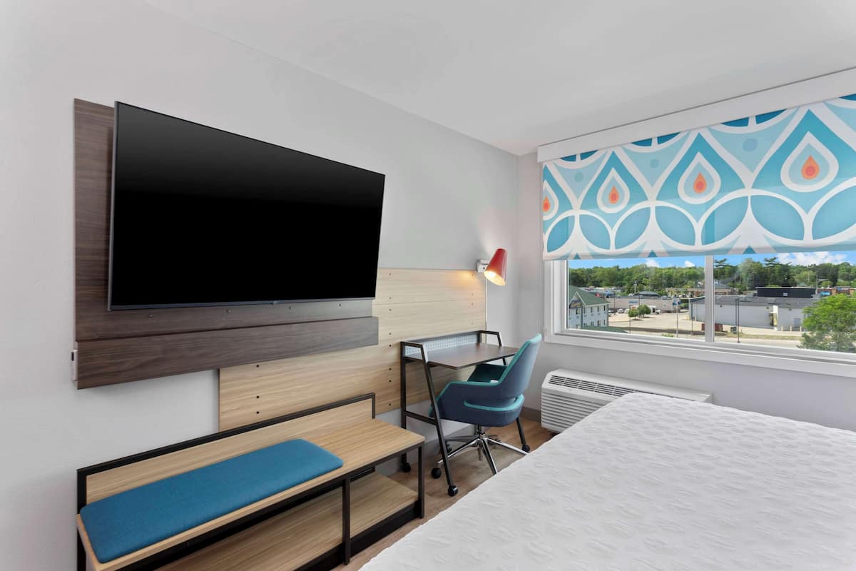 Habitaci ó d'hotel moderna amb televisió de pantalla plana penjada a la paret, escriptori amb cadira, una banca i una finestra gran amb vista a la ciutat.