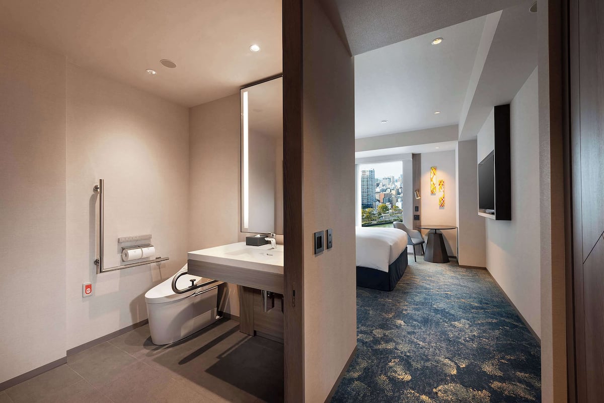 A modern hotel room with a bathroom, a bed, a TV, and a city view.