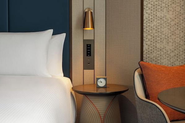 Habitación de hotel con cama, mesita de noche con un reloj, lámpara y una silla con cojín naranja.