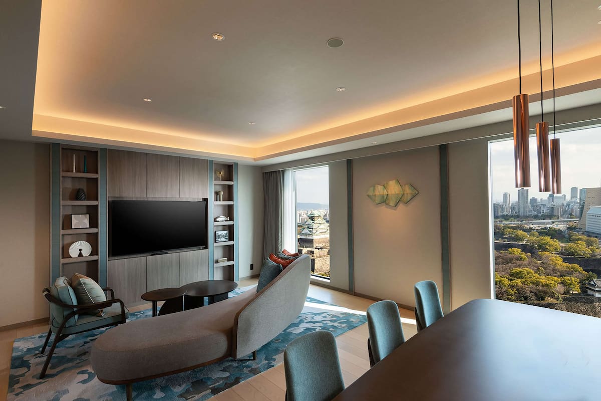 A modern living room with a flat-screen TV, comfortable seating, a dining area, and a large window offering a city view.