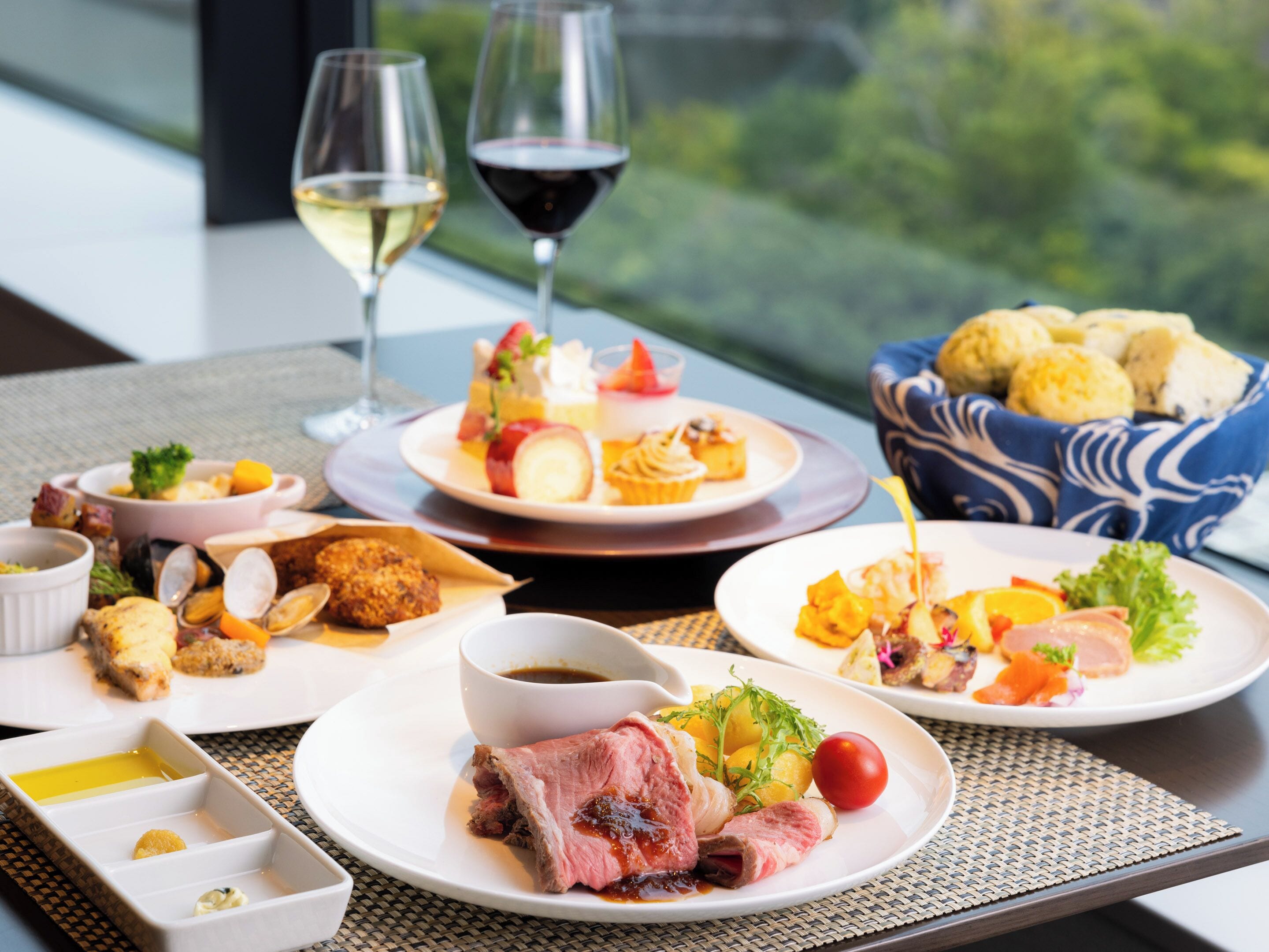 Una mesa dispuesta con diversos platos, incluyendo mariscos, carne y verduras, junto con copas de vino y vistas a la vegetación exterior.