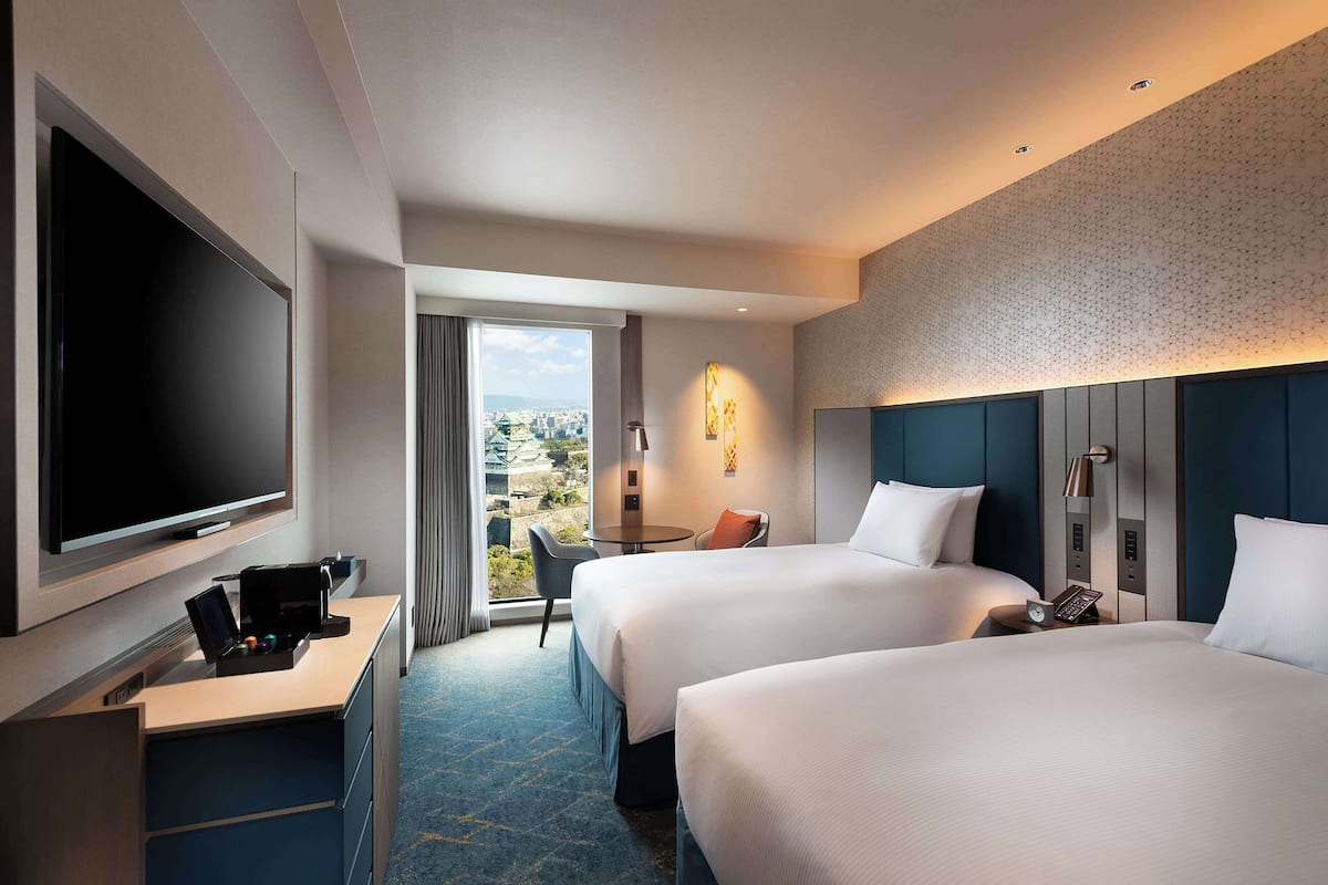 A hotel room with two beds, a TV, a desk with a phone, and a view of the city.