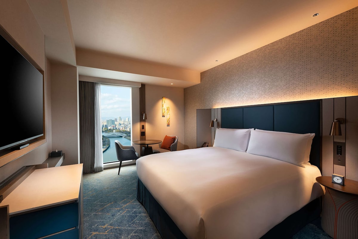 A hotel room with a large bed, a desk, a TV, and a view of a cityscape.