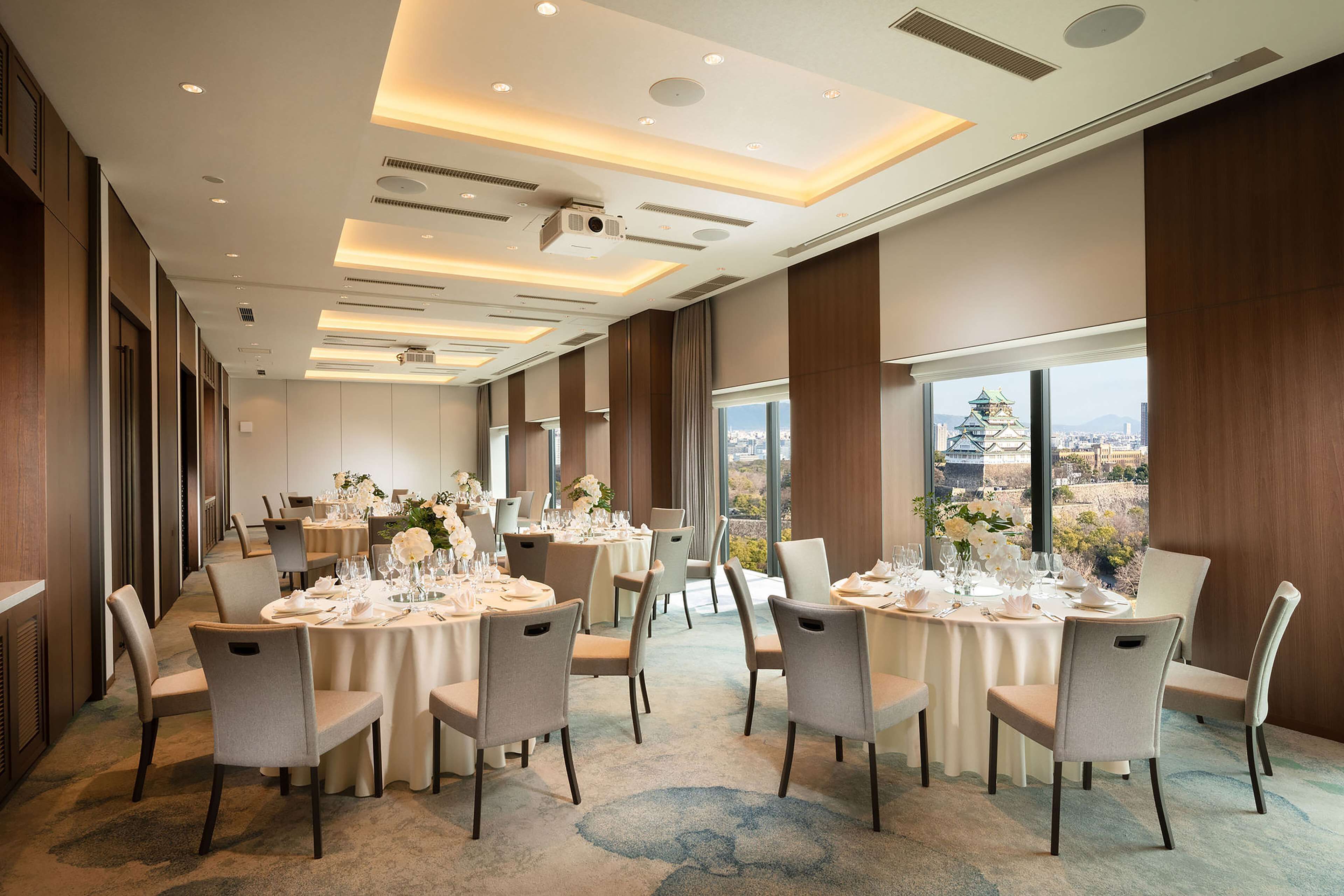 A hotel dining room with multiple round tables set for guests, large windows offering a city view, and a modern ceiling design.