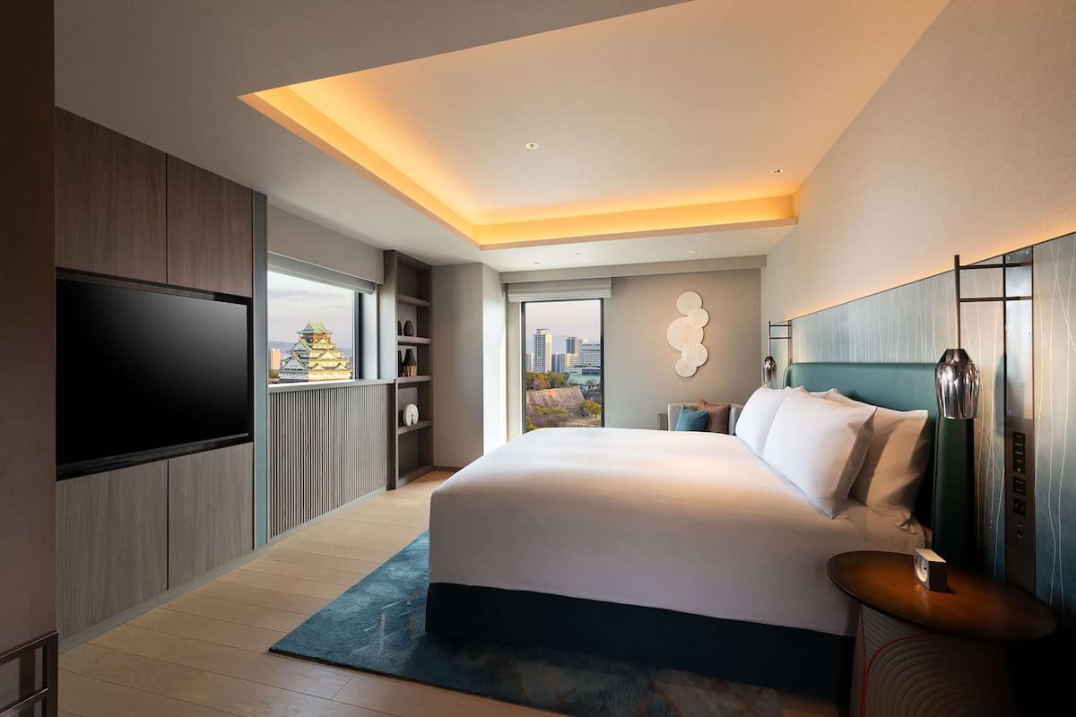 A modern hotel room with a large bed, a flat-screen TV, a city view, and a balcony.
