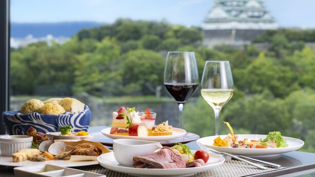 A table with various dishes and two glasses of wine, overlooking a lush green landscape and a traditional building. - DoubleTree by Hilton Osaka Castle (Osaka)
