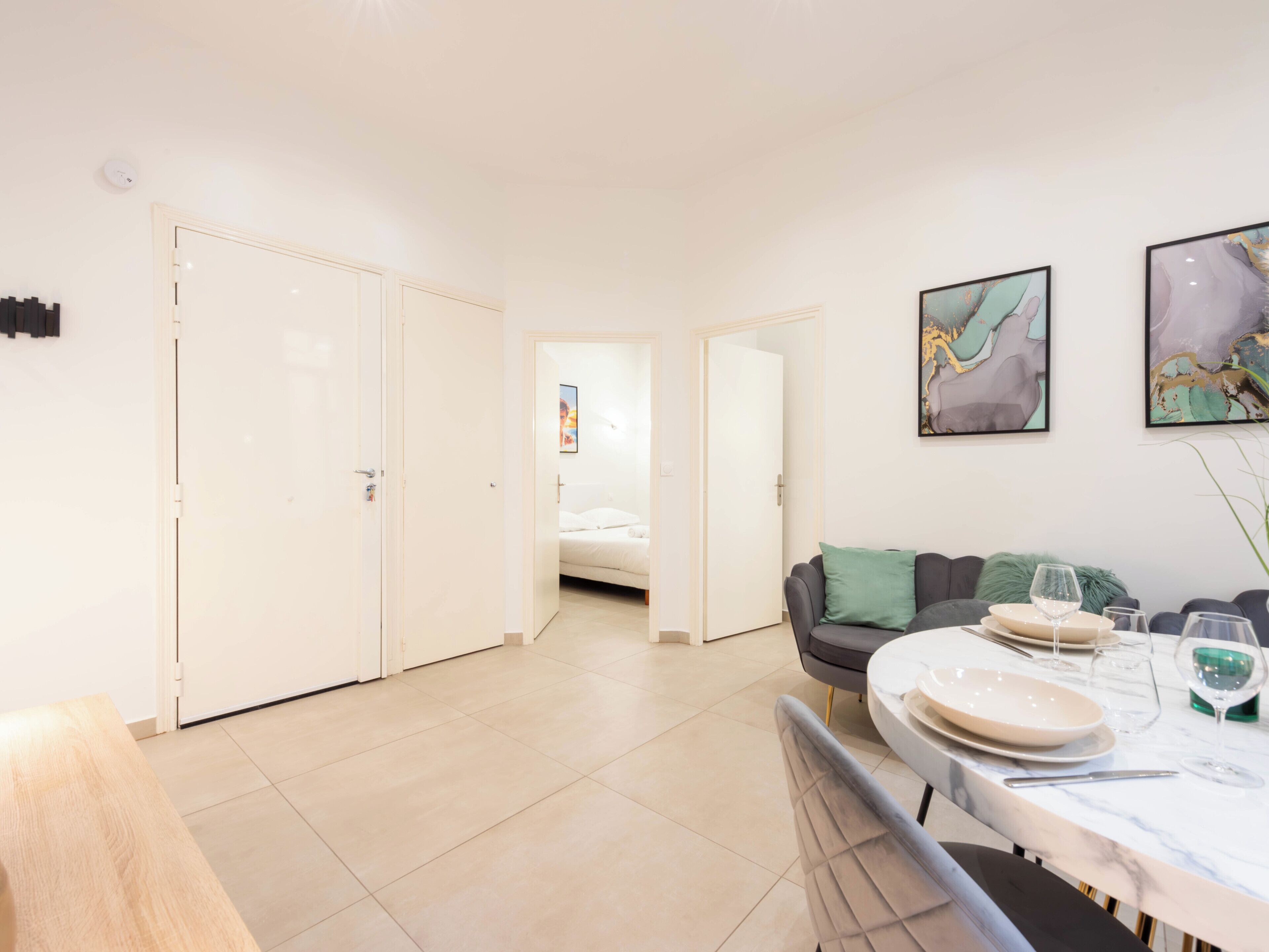 A modern, well-lit room with a dining table set for two, a sofa, and a bed in the background.