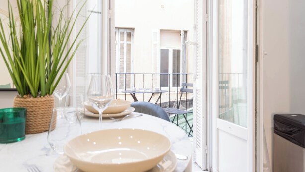 Un coin repas avec une table dressée pour deux personnes, une plante en pot et une vue sur un bâtiment par une porte ouverte.