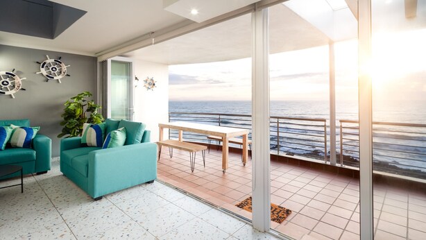 Un salon moderne avec des canapés de couleur turquoise, un banc en bois et une grande porte-fenêtre coulissante donnant sur un balcon avec vue sur la mer.