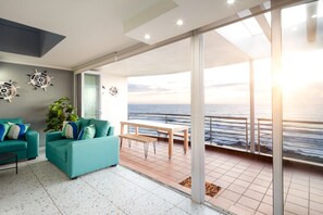 Un salon moderne avec des canapés de couleur turquoise, un banc en bois et une grande porte-fenêtre coulissante donnant sur un balcon avec vue sur la mer.