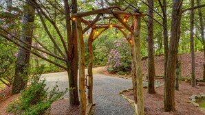 A wooden arbor with string lights, surrounded by trees and a gravel path. - The Cabin @ Possum Trot- A Rustic Chic Escape (Clayton)