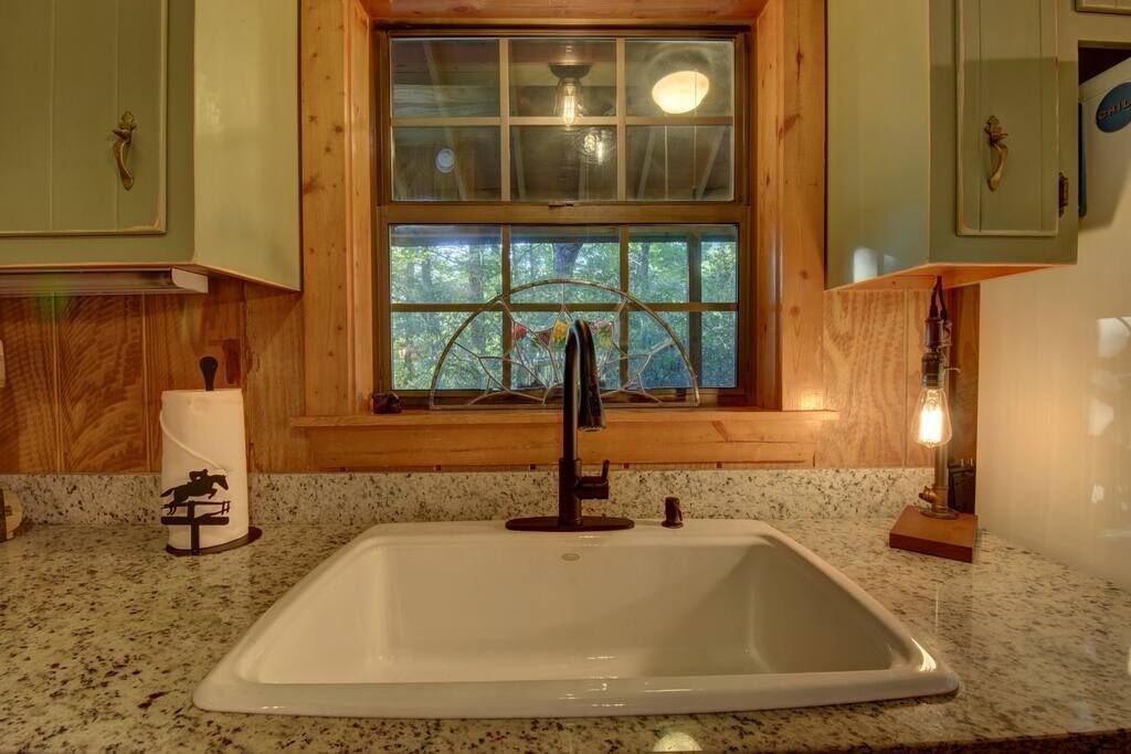 A kitchen with a white sink, wooden cabinets, and a window with a view of trees.