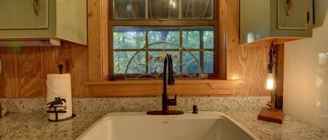 A kitchen with a white sink, wooden cabinets, and a window with a view of trees.