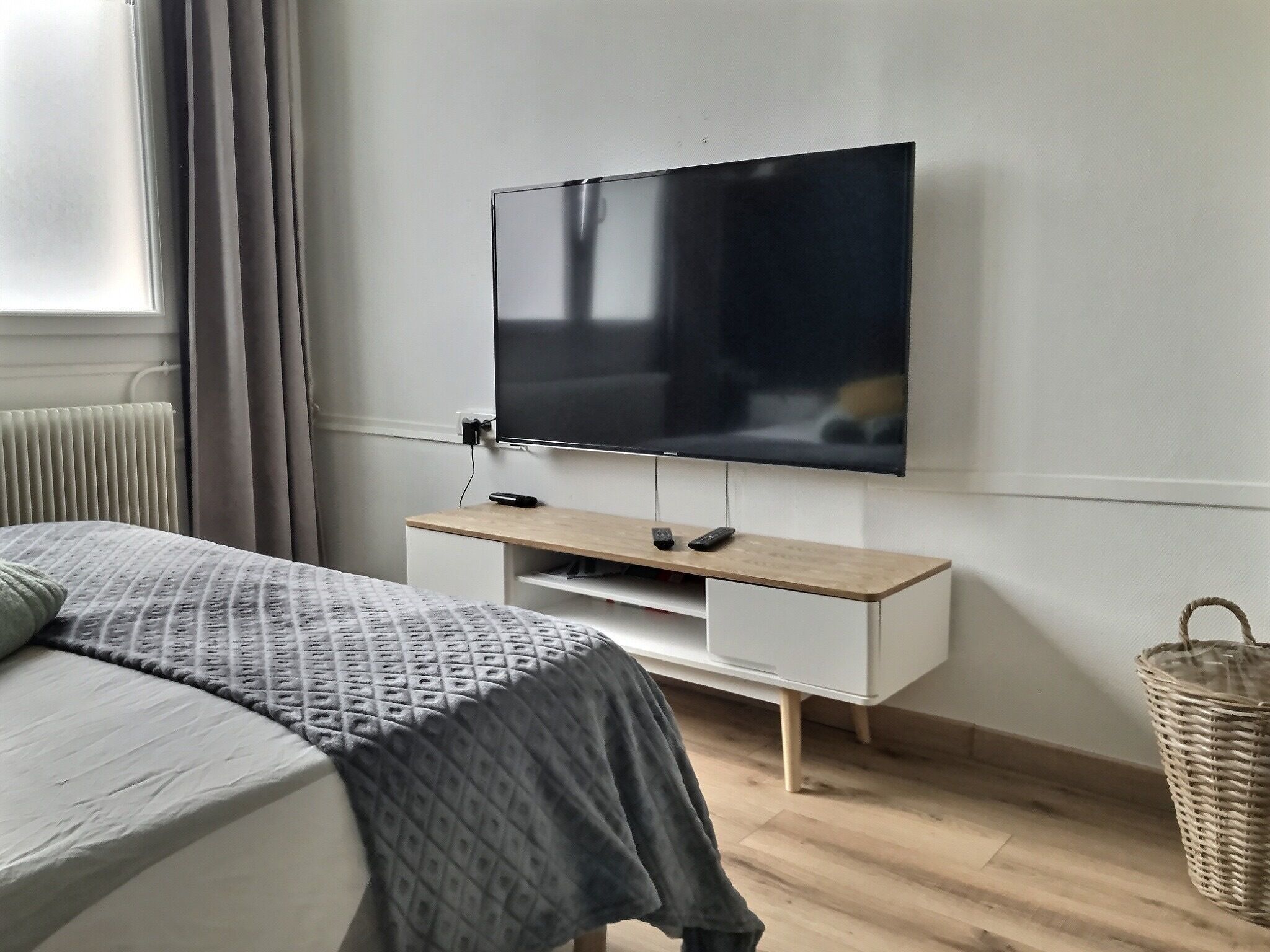Une chambre à coucher avec un lit, un téléviseur fixé au mur, une table de chevet et un panier en osier.