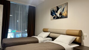 Une chambre d’hôtel avec deux lits, une grande fenêtre avec des rideaux, un tableau au mur et une table de chevet avec une lampe.
