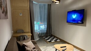 Un espace de vie compact comprenant un canapé, une petite table, un téléviseur, un meuble en bois et une vue sur l’extérieur à travers une fenêtre avec des rideaux.