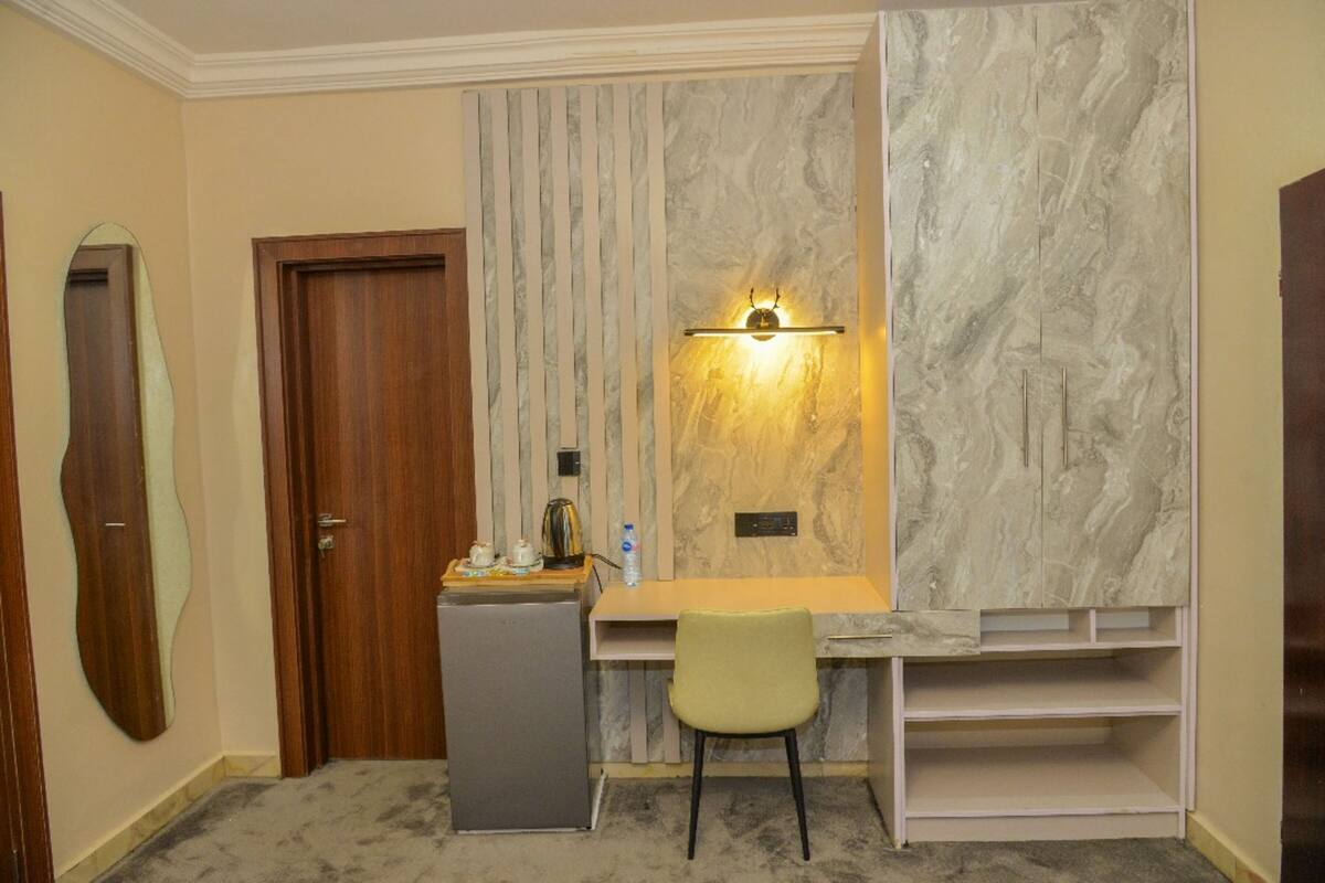 A compact room with a desk, a chair, a small refrigerator, and a wall-mounted light fixture.