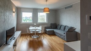 A modern living room with a grey sofa, a white dining table with chairs, a TV on a white stand, and a pendant light.