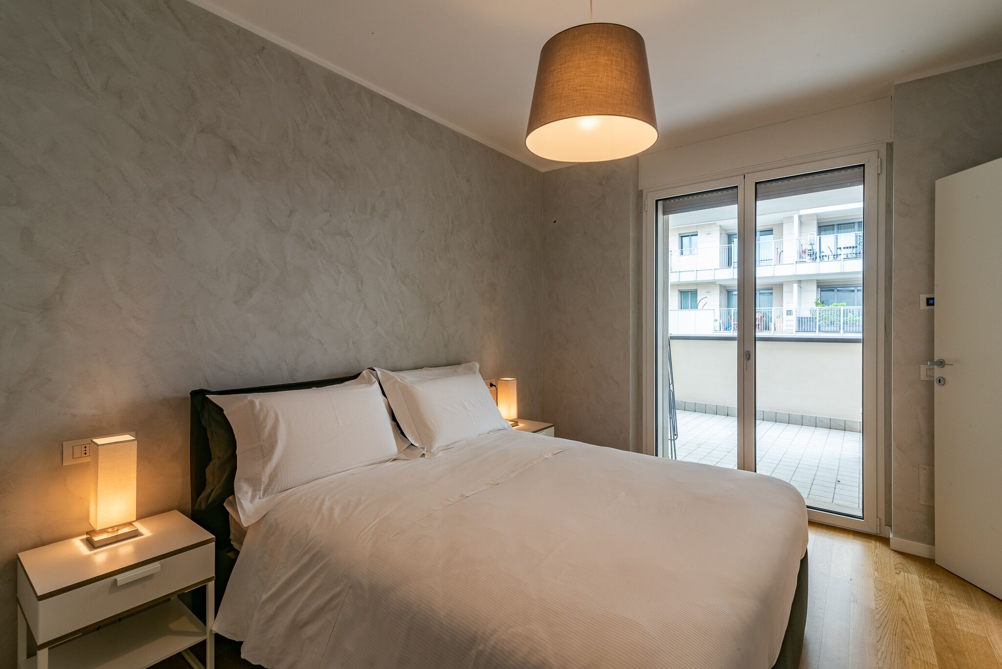 Une chambre à coucher avec un lit, des tables de chevet, une lampe et une vue sur un immeuble, visible à travers une porte vitrée.