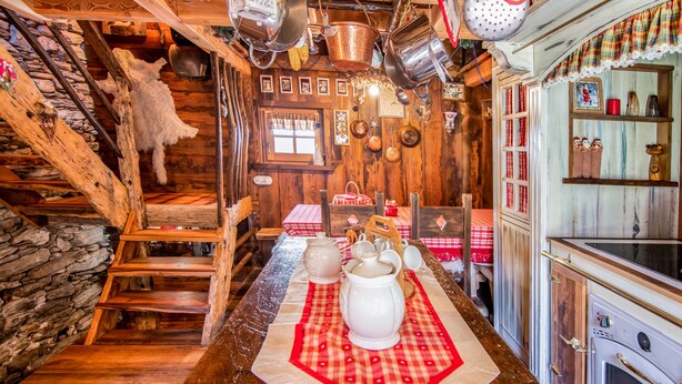 A rustic kitchen with wooden walls, a stone wall, and a wooden staircase. There is a table with a red and white checkered tablecloth and white ceramic pitchers. - 1 Bedroom Cabin in Isola (Isola)