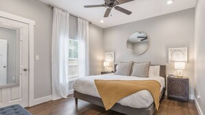 Un dormitorio con cama, ventilador de techo, mesita de noche con lámpara, espejo y puerta.