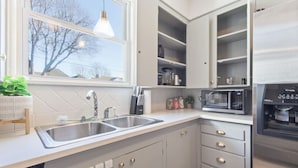 A modern kitchen with a double sink, built-in cabinets, a microwave, and a window with a view of trees. - Tulsa Vintage Chic Cottage |King Bed •Expo•TU•Dtwn (Tulsa)