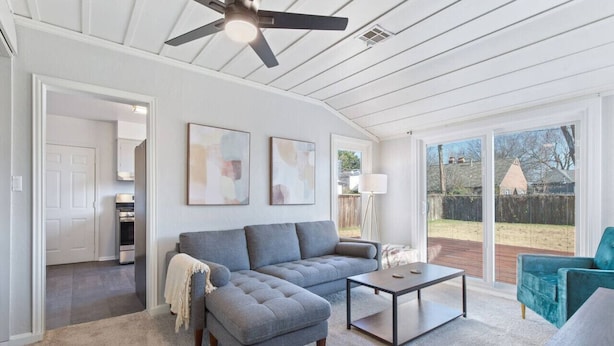 A spacious living room with a sectional sofa, a coffee table, and a ceiling fan. There are abstract paintings on the wall and a view of the backyard through large windows. - Tulsa Vintage Chic Cottage |King Bed •Expo•TU•Dtwn (Tulsa)