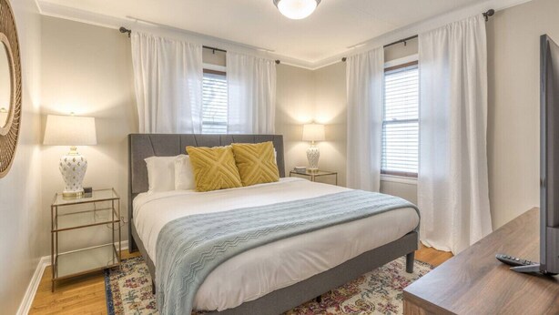 Quarto com cama, duas mesas de cabeceira com luminárias, televisão e janela com cortinas.
