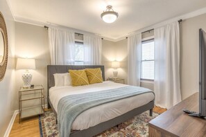 A bedroom with a bed, two bedside tables with lamps, a television, and a window with curtains. - OneCozyPlace Tulsa | King Bed • Expo•TU • Downtown (Tulsa)