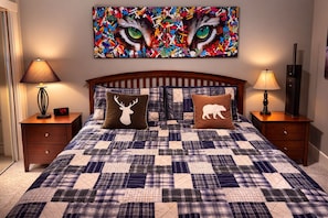 Quarto com colcha acolchoada, duas almofadas decorativas, mesa de cabeceira com abajur, relógio de parede e um quadro sobre a cama.