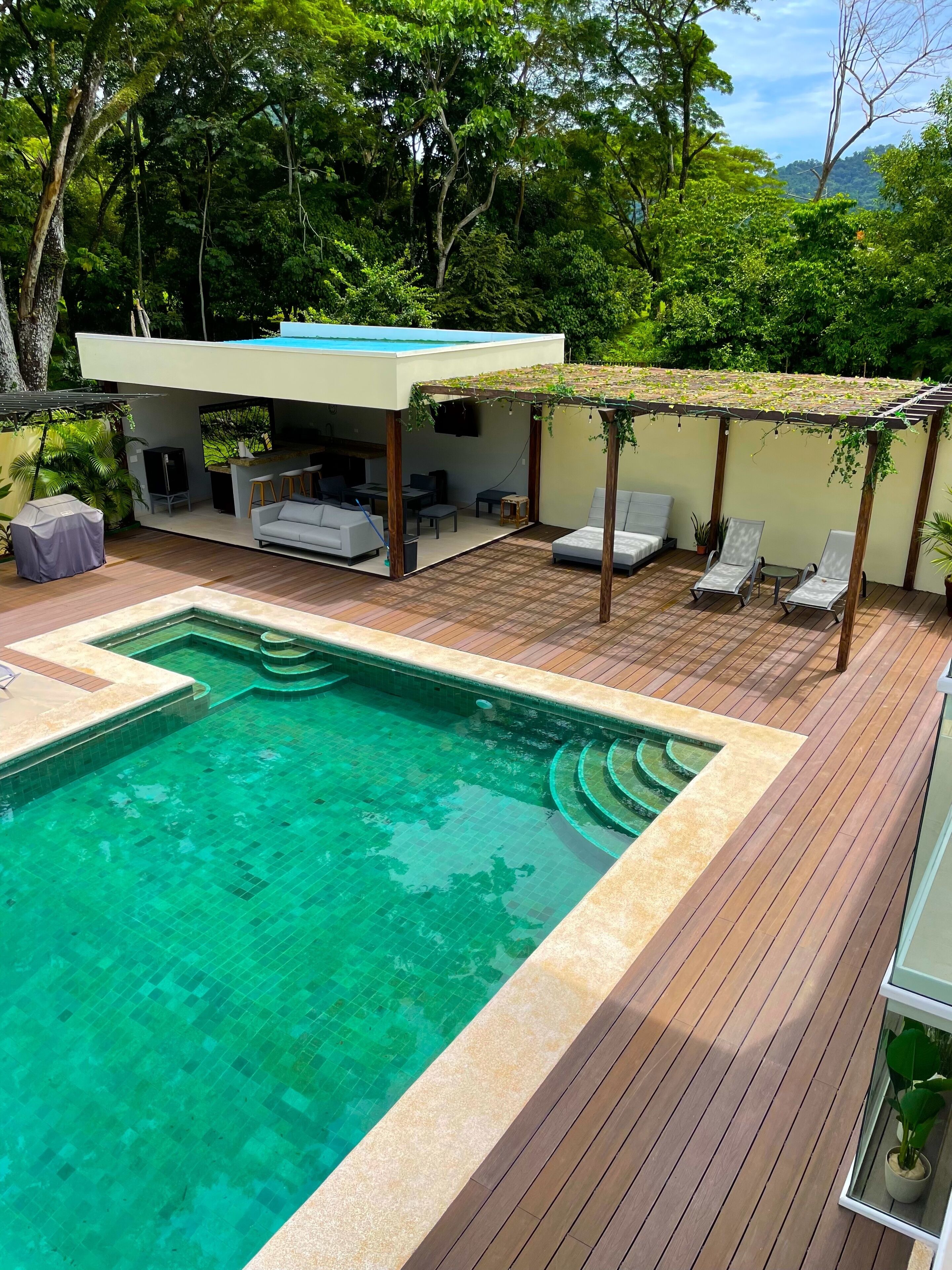 A swimming pool with a wooden deck, a covered seating area with a TV, and lush greenery in the background.