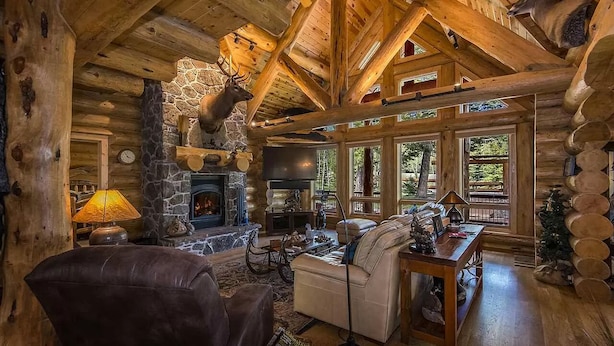 L’intérieur d’une cabane en rondins, avec une cheminée en pierre, des meubles en bois et une grande fenêtre donnant sur la forêt.