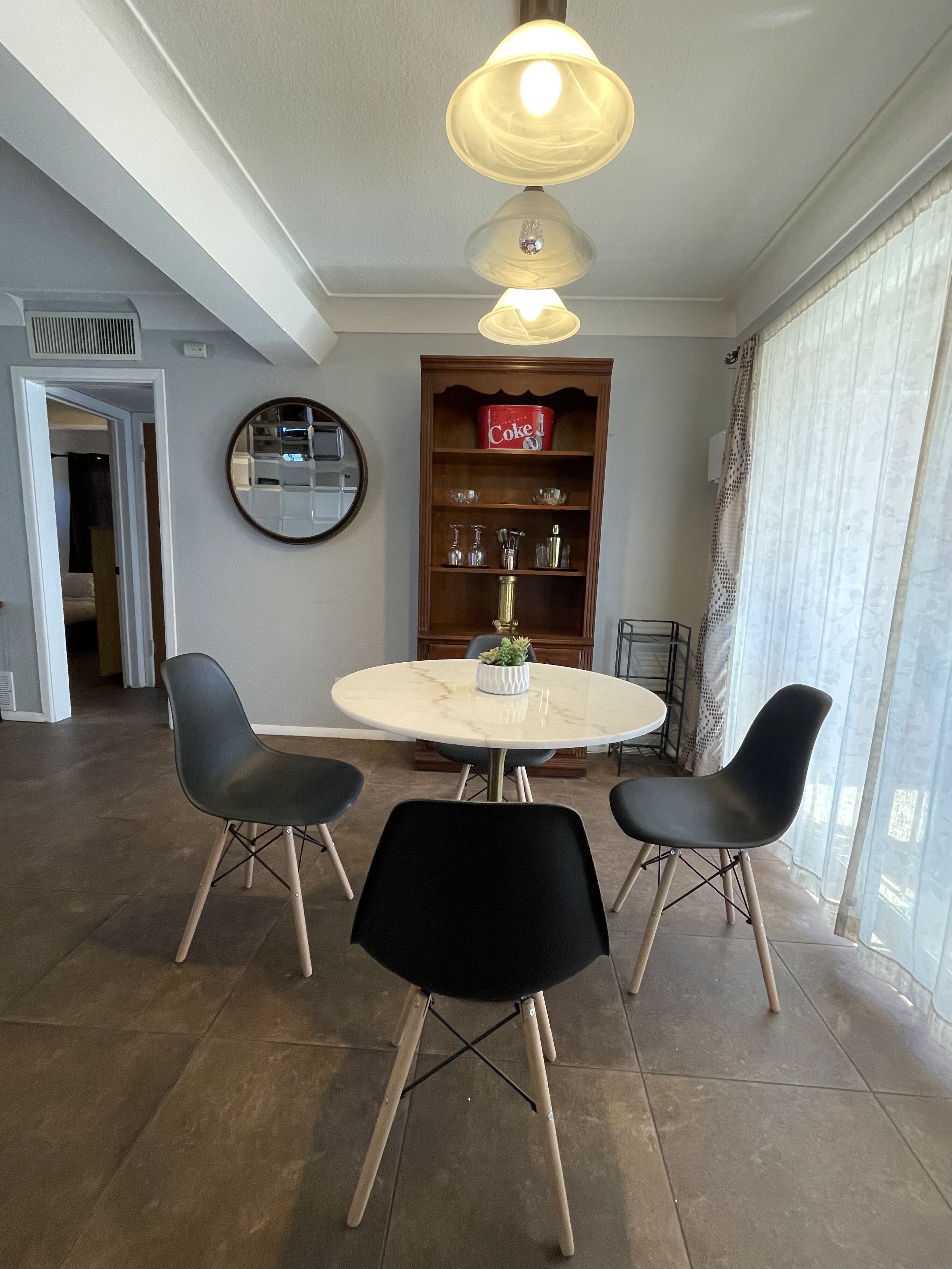 Un coin repas avec une table ronde, quatre chaises noires, un miroir, un réfrigérateur de la marque Coca-Cola et une fenêtre avec des rideaux.