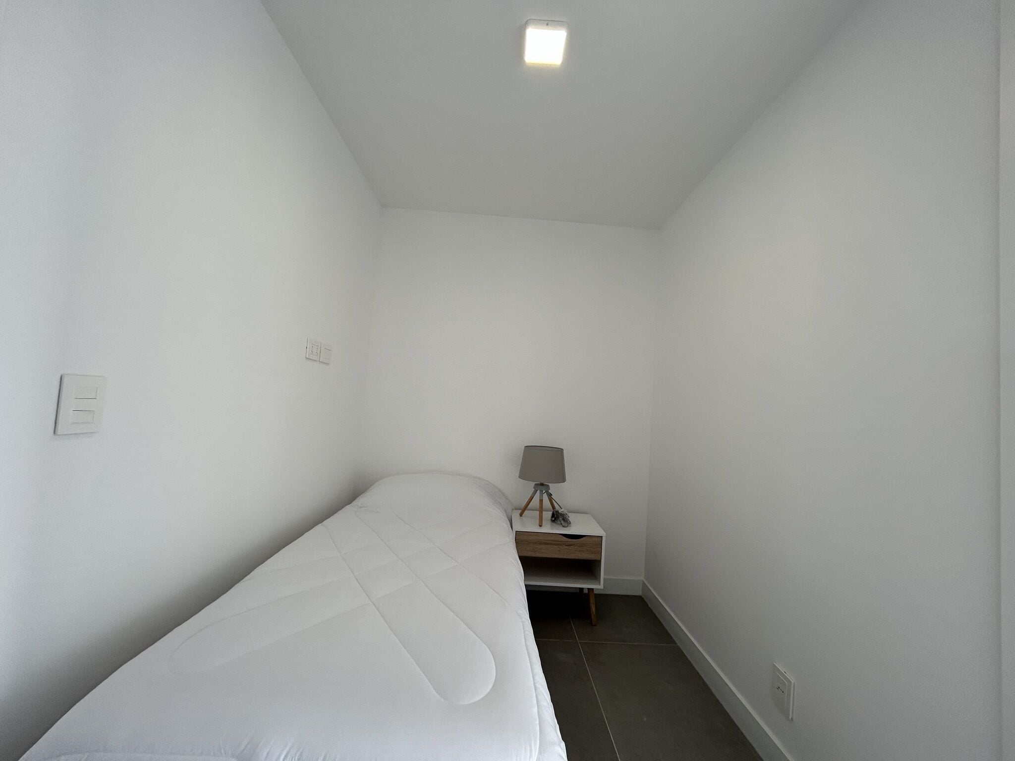 Une petite chambre blanche avec un lit simple, une table de chevet et une lampe.