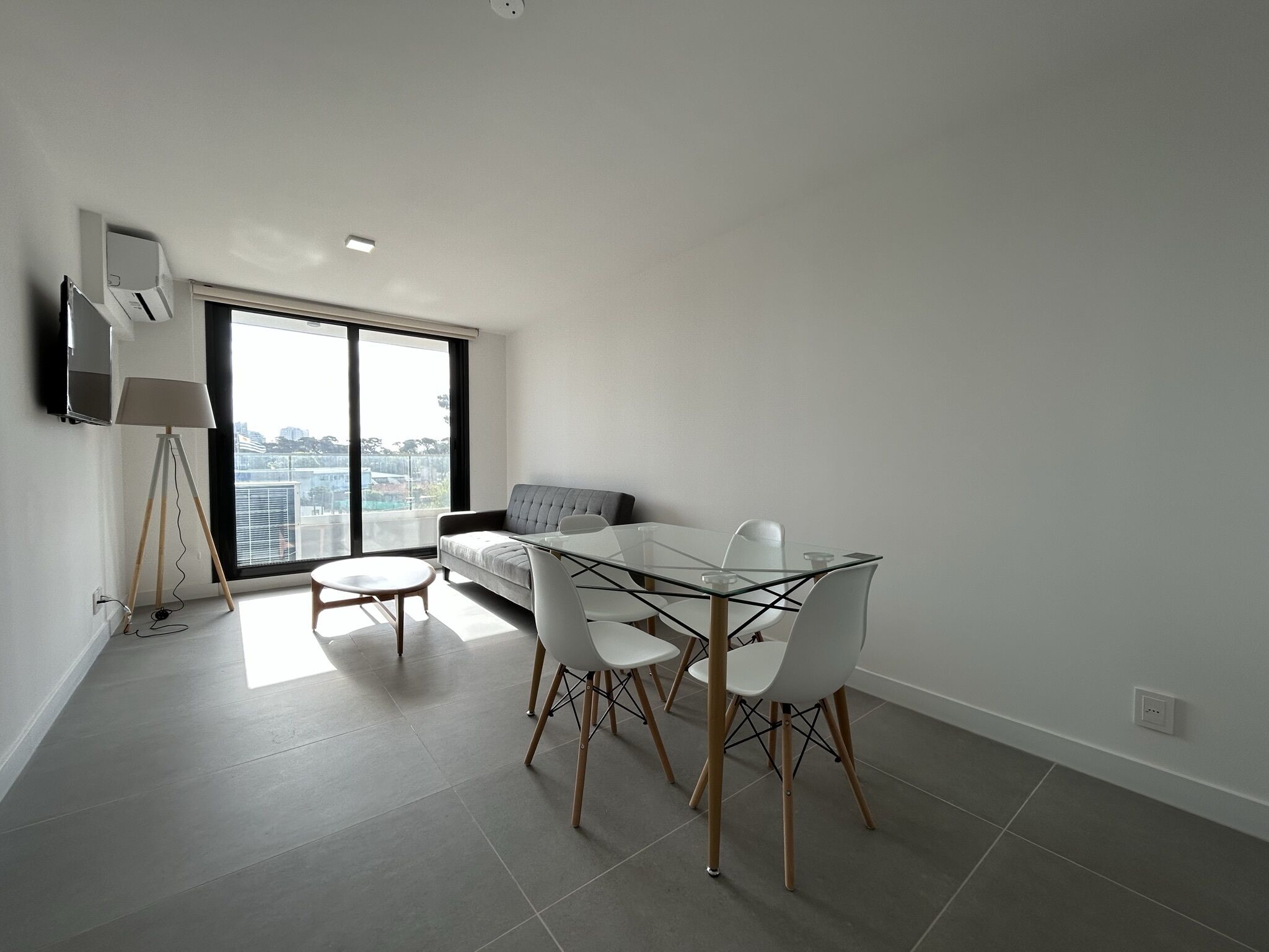 A modern dining area with a glass table and white chairs, a grey sofa, and a floor lamp.