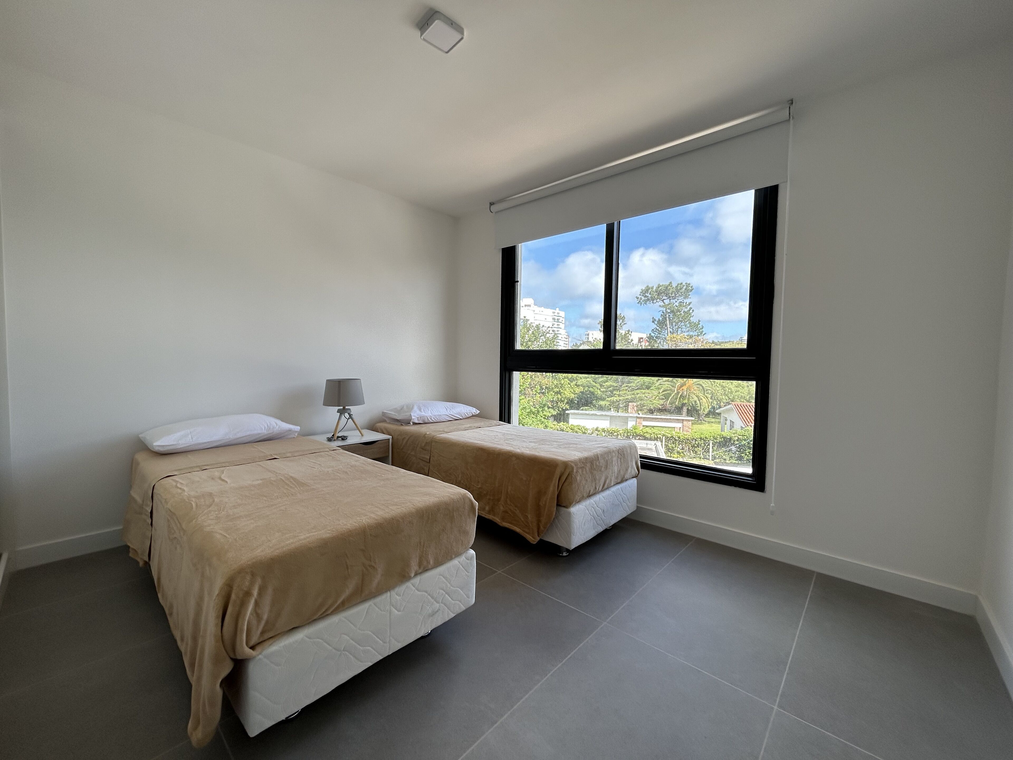 A bedroom with two beds, a nightstand, a lamp, and a large window with a view of buildings and greenery.