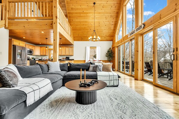 A spacious living room with a large sectional sofa, a round coffee table with candles, and a view of the outdoor deck. - Sleeps 16+ Hot Tub with Mountain View - 3Min to Sunday River Ski - Game Room

 (Newry)